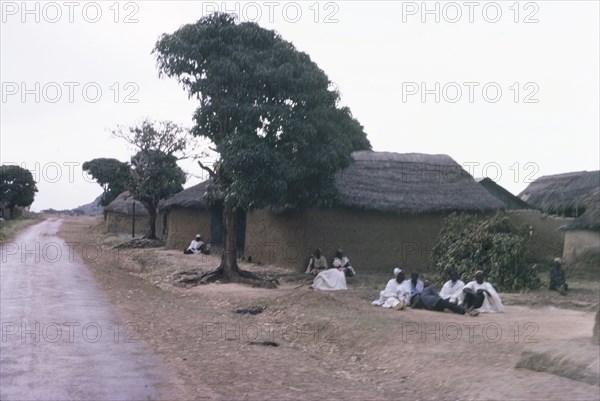 A Hausa village. Inhabitants of a Hausa village sit on the ground outside several square, mud-walled huts with thatched roofs. Nigeria, October 1963. Nigeria, Western Africa, Africa.