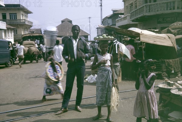 Market scene at Freetown, Sierra Leone. A woman balances a platter of fruit on her head, as she walks through a market in Freetown. A man in Western-style clothes and winklepicker shoes stands behind her, posing for the camera. Freetown, Sierra Leone, 12 February 1964. Freetown, West (Sierra Leone), Sierra Leone, Western Africa, Africa.