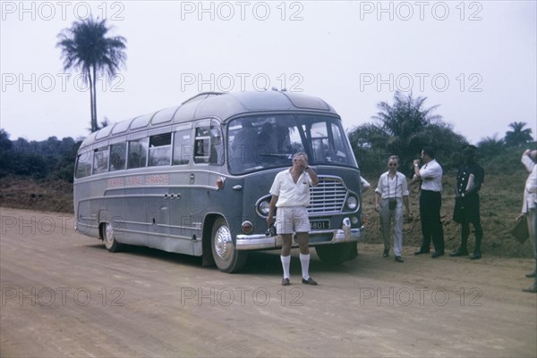 A broken-down bus. Inconvenienced European passengers stand on a roadside beside a broken-down bus, whilst a uniformed African official looks on. Freetown, Sierra Leone, 1964. Freetown, West (Sierra Leone), Sierra Leone, Western Africa, Africa.
