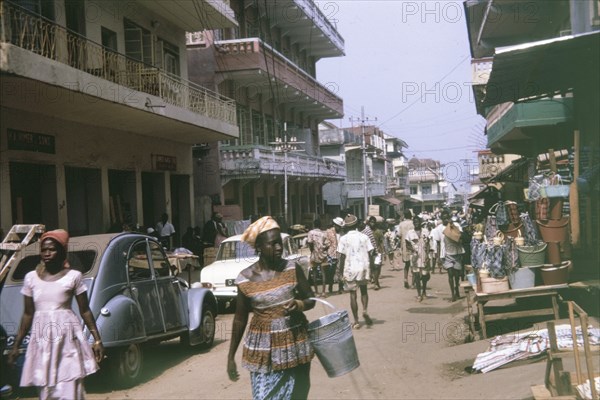Busy urban street in Freetown. Pedestrians and road traffic crowd a busy urban street in Freetown, which is flanked by multi-storey buildings with balconies. Freetown, Sierra Leone, February 1964. Freetown, West (Sierra Leone), Sierra Leone, Western Africa, Africa.