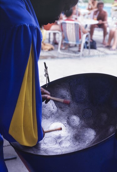 Barbadian musician playing steel drum. A Barbadian musician bows his head over a steel drum as he plays for an audience of tourists lounging beside a swimming pool. Barbados, circa 1975. Barbados, Caribbean, North America .