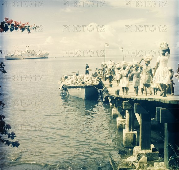 Maritime trade, Dominica. A group of women walk along a wooden pier carrying sacks on their heads, which they load into a motor boat waiting in harbour. A luxury liner is moored offshore, possibly waiting to receive the supplies. Dominica, circa 1975. Dominica, Caribbean, North America .