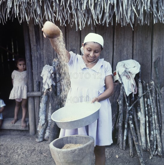 Separating the grain from the husk. A Ketchi woman pours rice from one bowl into another, separating the grain from the husk before grinding the rice inside a large pestle and mortar. Belize, circa 1975. Belize, Central America, North America .