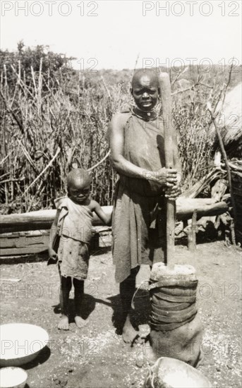 Preparing food outdoors, Kenya. A shy young child clings to his mother as she prepares food outdoors using a large pestle and mortar. Rift Valley, Kenya, circa 1933., Rift Valley, Kenya, Eastern Africa, Africa.