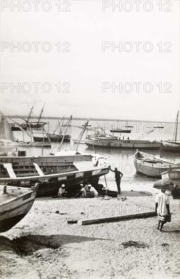 Dry-docked boats in Lamu harbour. A group of men sit on a beach beside a dry-docked boat in Lamu harbour. Lamu, Kenya, circa 1947. Lamu, Coast, Kenya, Eastern Africa, Africa.