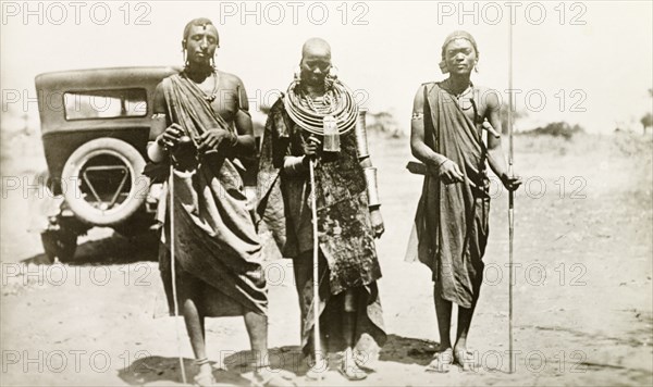 Maasai portrait. Portrait of two Maasai men and a Maasai woman. They stand in a line on a rural road, each holding a staff and wearing traditional dress and jewellery. British East Africa (Kenya), circa 1912. Kenya, Eastern Africa, Africa.