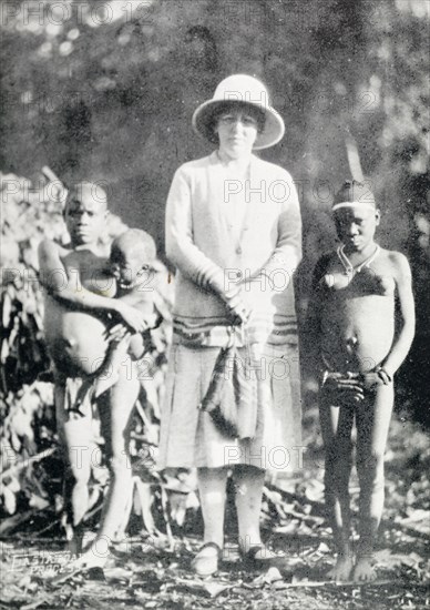 A British woman poses with "pigmy women". A British woman poses for a portrait in between two semi-naked African woman, who are identified in an original caption as "full grown pigmy (sic) women". British East Africa (Kenya), circa 1912. Aba, Abd Allah, Kenya, Eastern Africa, Africa.