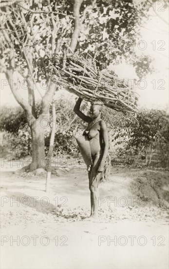 Fuel gatherer'. Profile shot of a semi-naked African woman. She stands, balancing a large bundle of firewood on her head whilst carrying a baby strapped to her back. British East Africa (Kenya), circa 1912. Kenya, Eastern Africa, Africa.