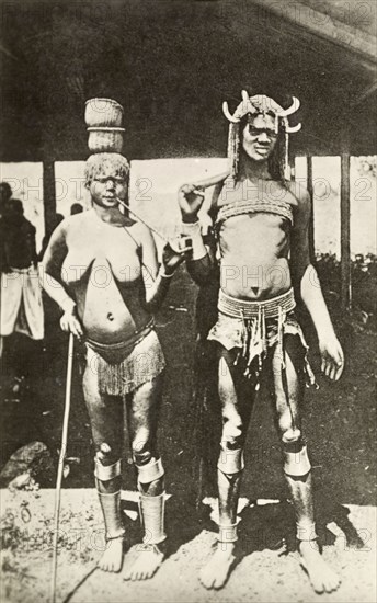 Portrait of a Kavirondo couple. Full-length portrait of a semi-naked Kavirondo (Luo) couple. Dressed in a grass skirt and anklets, the woman smokes a long pipe and leans on a stick. The man wears a chest strap and loincloth with his hair braided and decorated with horn ornaments. British East Africa (Kenya), circa 1912. Kenya, Eastern Africa, Africa.
