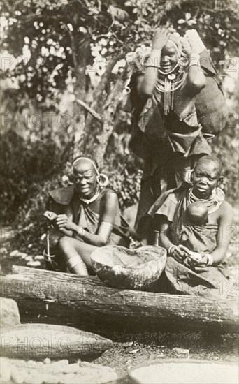 Three Kikuyu women. Portrait of three Kikuyu women wearing traditional dress and jewellery including masses of hooped earrings and necklaces. Two of the women prepare food as they sit on the ground, whilst the third stands, carrying a large bundle of firewood on her back. British East Africa (Kenya), circa 1912. Kenya, Eastern Africa, Africa.