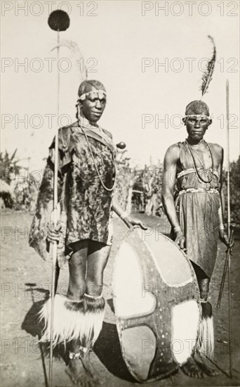Two Maasai warriors. Full-length portrait of two Maasai warriors in traditional dress, holding spears and a patterned shield. British East Africa (Kenya), circa 1912. Kenya, Eastern Africa, Africa.