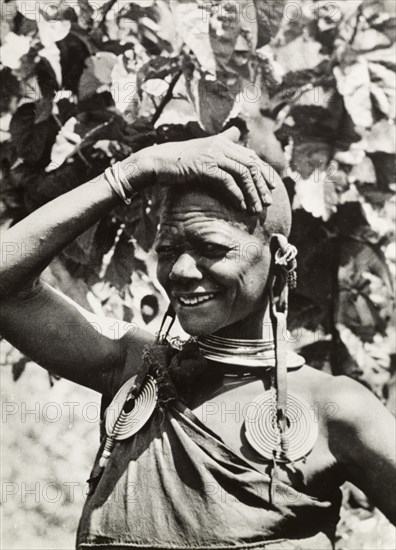 One of Chief Murigo's 19 wives'. Portrait of a Kikuyu woman, wearing several items of jewellery including a pair of heavy, ornate earrings and a necklace. She is identified in an original caption as ?One of Chief Murigo's 19 wives?, her marriage status indicated by her shaven head. South Nyeri, Kenya, 1936. Nyeri, Central (Kenya), Kenya, Eastern Africa, Africa.