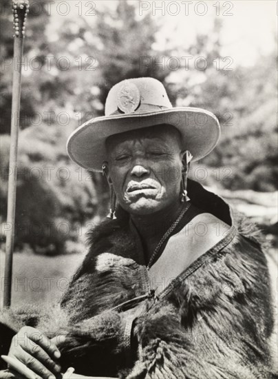 Chief Murigo. Portrait of Chief Murigo wearing traditional Kikuyu jewellery and holding a staff. He wears a Western-style, wide-brimmed hat with a badge depicting a lion, and an animal skin wrapped around his shoulders. Near Kakamega, Kenya, 1936. Kakamega, West (Kenya), Kenya, Eastern Africa, Africa.
