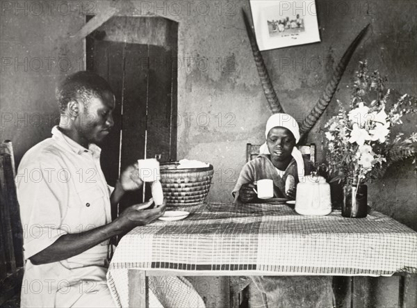 A Kikuyu couple in a Western-style home. A Kikuyu employee of the government's Agricultural Department sits at a table drinking tea with his wife in their new Western-style home. An original caption comments that this new style of house was "square instead of round, with windows and doors". Kenya, 1936. Kenya, Eastern Africa, Africa.