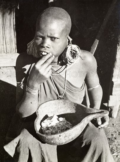Kikuyu woman eating maize porridge. A Kikuyu woman sits on the ground eating a meal of maize porridge from a hollowed-out gourd. Her head is shaven, indicating her status as a married woman, and she wears masses of hooped earrings. South Nyeri, Kenya, 1936. Nyeri, Central (Kenya), Kenya, Eastern Africa, Africa.