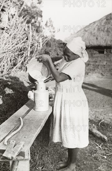 Kikuyu woman pouring milk. A Kikuyu woman, wearing a Western-style dress and headscarf, pours milk from a bucket into a milk churn on an outdoor workbench. South Nyeri, Kenya, 1936. Nyeri, Central (Kenya), Kenya, Eastern Africa, Africa.