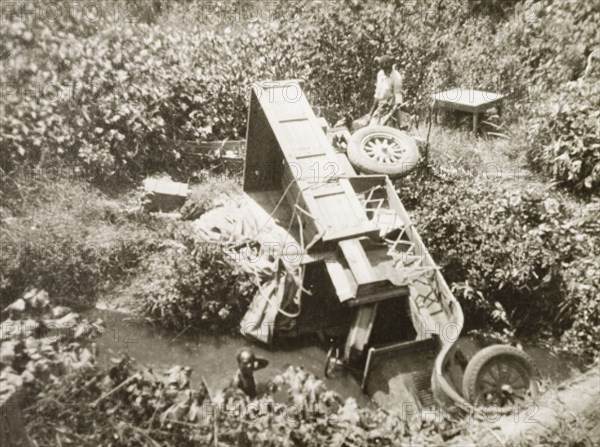 Sometimes things go wrong'. The wreckage of a truck lies in a stream by the side of a road. Kenya, circa 1925. Kenya, Eastern Africa, Africa.