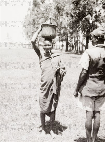 A Luo woman smoking. Portrait of a Luo woman wearing a simple robe. She balances a basket on her head and smokes a long clay pipe. Kakamega, Kenya, 1933. Kakamega, West (Kenya), Kenya, Eastern Africa, Africa.