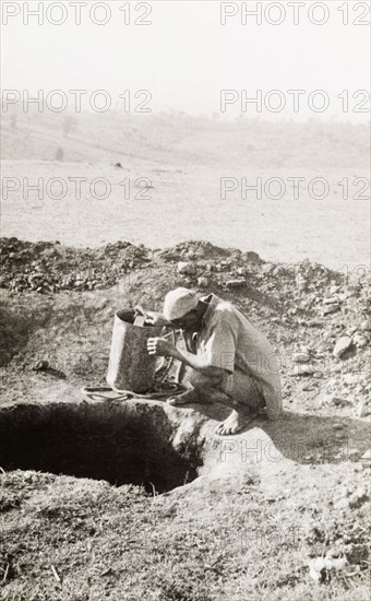 Exploratory mine shaft at the Kakamega goldfields. A labourer squats at the opening to a mine shaft on the Kakamega goldfields. According to an original caption, this was "an exploratory pit sunk to strike the (gold) reef and discover its direction". Kakamega, Kenya, 1933. Kakamega, West (Kenya), Kenya, Eastern Africa, Africa.