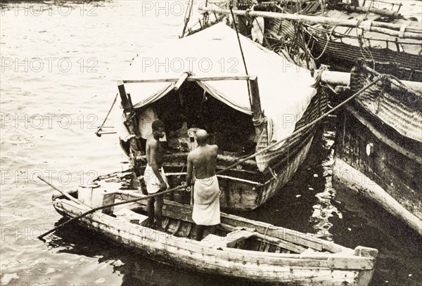Attending to cargo at Old Mombasa harbour. Two men stand in a canoe, attending to cargo stored beneath the canvas shelter of a dhow moored in Old Mombasa harbour. Mombasa, Kenya, 1933. Mombasa, Coast, Kenya, Eastern Africa, Africa.