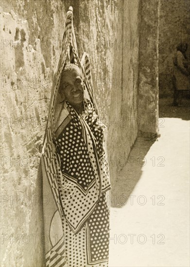 Wrapped in a swathe of fabric. An elderly person leans against a wall in Lamu, wrapped in a swathe of patterned fabric, possibly a flag. Lamu, Kenya, 1947. Lamu, Coast, Kenya, Eastern Africa, Africa.