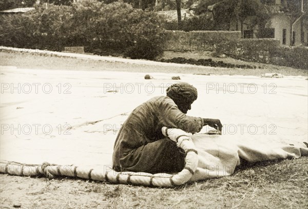 A sail maker repairs a dhow sail. A sail maker repairs a dhow sail on an open patch of ground below Fort Jesus. Mombasa, Kenya, 1933. Mombasa, Coast, Kenya, Eastern Africa, Africa.
