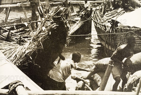 Unloading cargo at Old Mombasa harbour. Men unload cargo from beneath the thatched canopy of a dhow moored in Old Mombasa harbour. Mombasa, Kenya, 1933. Mombasa, Coast, Kenya, Eastern Africa, Africa.