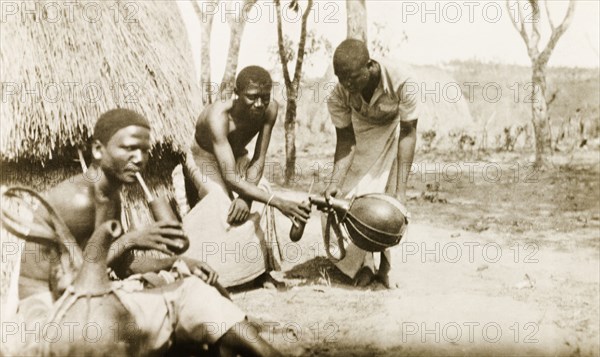 Pouring drinks from a gourd. A Kenyan man pours his two friends a drink from a gourd, one of whom sits in front of a thatched hut, drinking through a thick straw. British East Africa (Kenya), circa 1912. Kenya, Eastern Africa, Africa.
