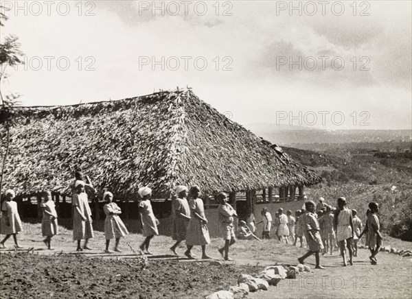 Kikuyu schoolchildren. A group of Kikuyu schoolchildren walk in single file towards a thatched roof school building in Nyeri. An original caption comments: "The modern world starts to break the mould of Kikuyu society, with Western (literary) education". Nyeri, Kenya, 1936. Nyeri, Central (Kenya), Kenya, Eastern Africa, Africa.