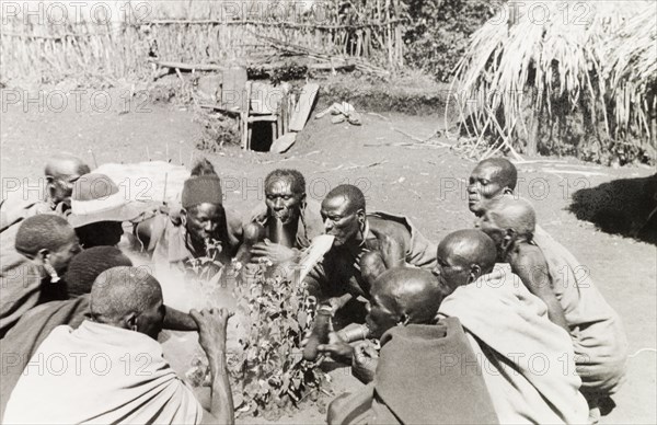 Kikuyu good fortune ceremony. A group of Kikuyu men gather in a circle, performing a ceremony to bring good fortune to anyone embarking on a journey. An original caption comments: "(A) witchdoctor collects herbs... from all the paths leading to the village, and spits on them (to bring) good luck". Nyeri, Kenya, 1936. Nyeri, Central (Kenya), Kenya, Eastern Africa, Africa.