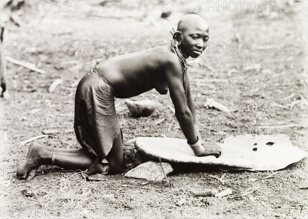 Grinding millet. A Kikuyu woman, naked from the waist up, kneels on the ground as she grinds millet on a flat board. Probably Nyeri, Kenya, circa 1936. Nyeri, Central (Kenya), Kenya, Eastern Africa, Africa.