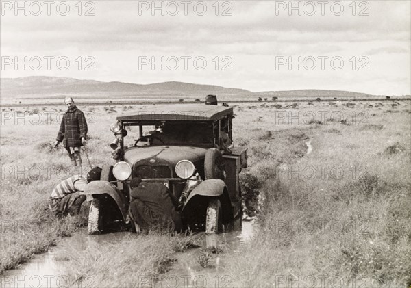 Stuck in the mud. Several men attend to a car, which is stuck in the mud on a flooded country track. North Tanganyika Territory (Tanzania), circa 1933. Tanzania, Eastern Africa, Africa.