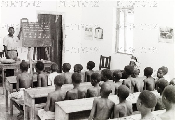Reading lesson at a Kenyan school. Schoolchildren attend a reading lesson at a school in Kenya. Their teacher stands beside a blackboard inscribed with vowel letters and their associated phonetics: perhaps an indication that this is an English lesson. Coastal Region, Kenya, 11 February 1947., Coast, Kenya, Eastern Africa, Africa.