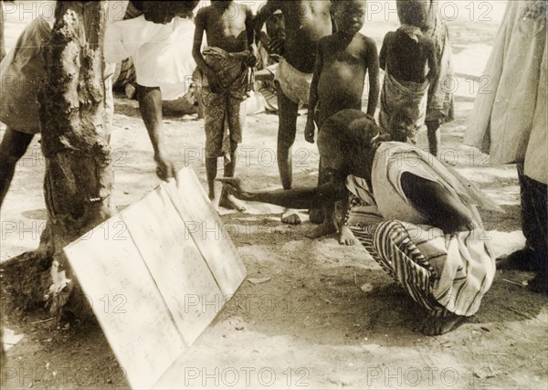 Adult literacy campaign, Gold Coast. A student teacher helps a man to read from a board propped up against the base of a tree during a literacy class: part of a mass literacy campaign launched by the British government in 1951 to improve literacy skills amongst African adults, both in their own languages and in English. Northern Territories, Gold Coast (Northern Ghana), circa 1951., North (Ghana), Ghana, Western Africa, Africa.