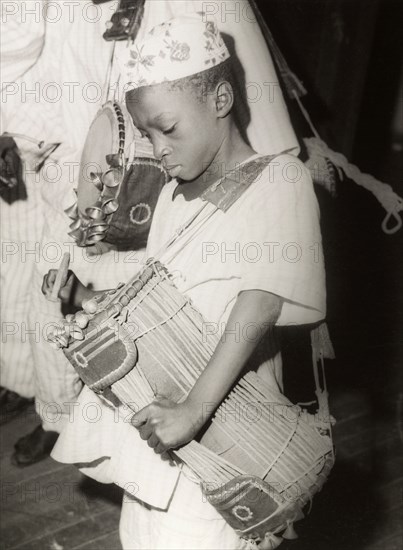 Child playing a 'dundun' drum. A child plays a 'dundun' (talking drum), a double-headed drum shaped like an hourglass with a number of cords running the length of the shell. These cords allow the drummer to adjust the tension of the instrument, resulting in a change of pitch that imitates the sounds of language. Nigeria, circa 1965. Nigeria, Western Africa, Africa.