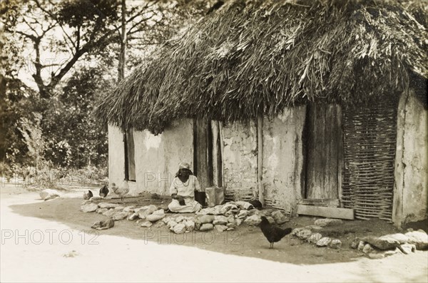 Woman and baby under the eaves. A woman and baby sit in the shade beneath the thatched eaves of a wattle and daub hut. Jamaica, circa 1925. Jamaica, Caribbean, North America .