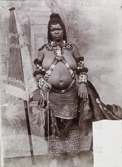 Portrait of a young African woman. Studio portrait of young African woman, possibly a bride, in traditional dress. The woman stands against a backcloth painted with an ornate gate and foliage. A large floral and lace parasol serves as a prop. Nigeria, circa 1925. Nigeria, Western Africa, Africa.