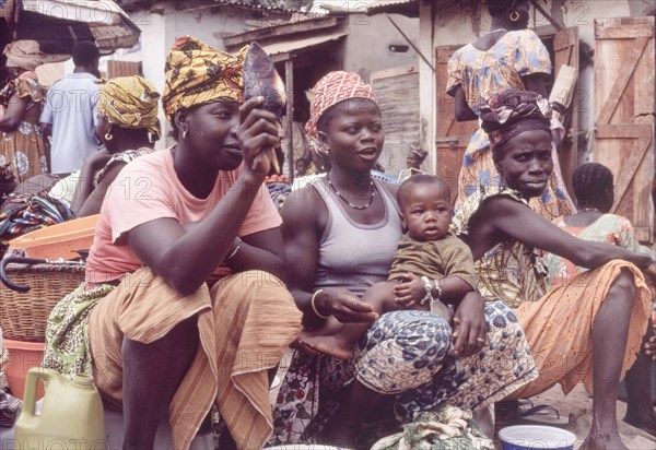 Female traders at Banjul market. Female traders sell food produce at a bustling outdoor market in Banjul. One of the women balances a baby on her knee, whilst another holds up a smoked fish for the camera. Banjul, Gambia, circa 1985. Banjul, Banjul City, Gambia, Western Africa, Africa.