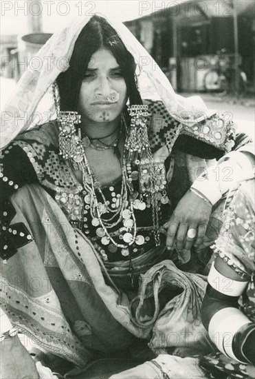 A woman wearing elaborate Indian jewellery. Seated portrait of an Indian woman, identified in an original caption as a 'gypsy'. Her head is loosely covered with a veil and she wears elaborate jewellery including a nose ring, necklaces and ornate hair ornaments. Her traditional clothing is embroidered with patterns and decorated with mirrorwork. Mapusa, Goa, India, circa 1978. Mapusa, Goa, India, Southern Asia, Asia.