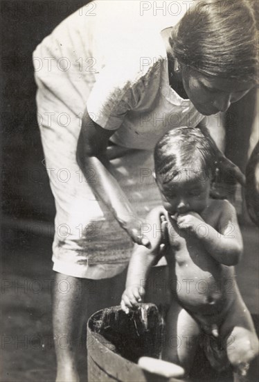 A European woman washes her child. A European woman bends over to wash her young child in a washtub outdoors. Trinidad and Tobago, circa 1931. Trinidad and Tobago, Caribbean, North America .