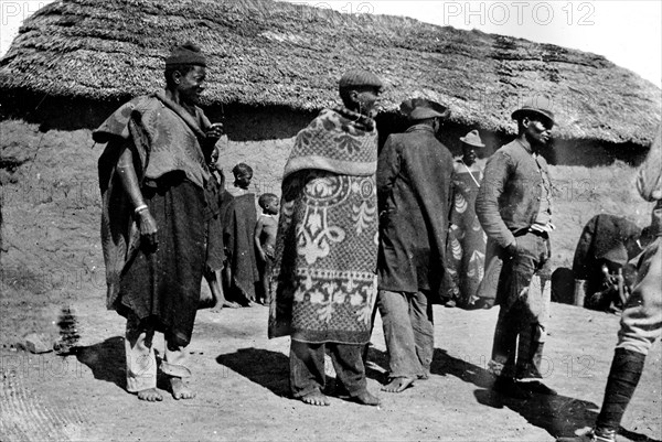 African men outside a hut. African men, possibly members of a Bantu-speaking tribe, mill about in front of a mud-walled thatched hut. They are barefoot and wrapped in swathes of patterned fabric. South Africa, circa 1901. South Africa, Southern Africa, Africa.