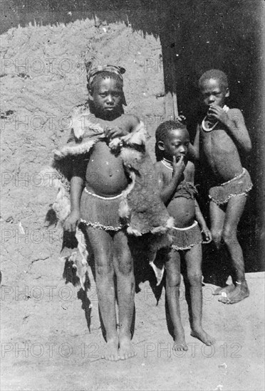 Three African boys. Three young boys, possibly members of a Bantu-speaking tribe, pose for the camera in front of a mud-walled hut. They wear beaded skirts and necklaces, the eldest wrapped in an animal skin. South Africa, circa 1901. South Africa, Southern Africa, Africa.