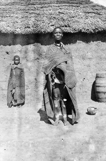 An African woman and child. An African woman and child, possibly members of a Bantu-speaking tribe, pose for a portrait outside a mud-walled thatched hut. The woman is wrapped in a swathe of patterned fabric and wears jewellery including necklace and anklets. South Africa, circa 1901. South Africa, Southern Africa, Africa.