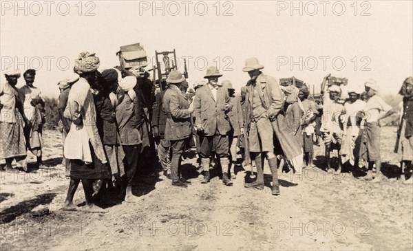 Colonial hunting party with 'shikaris'. Sir Henry Staveley Lawrence, Acting Governor of Bombay, chats with companions as they prepare to embark on a hunting expedition. A large party of 'shikaris' (professional hunters) and porters assemble behind them: some carrying chairs, others hunting equipment. Drigh, Karachi, Sind, India (Sindh, Pakistan), 1926. Karachi, Sindh, Pakistan, Southern Asia, Asia.