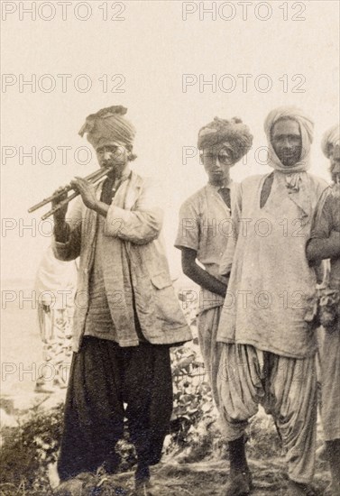Musician playing a 'satara'. A musician plays a 'satara' (Indian dual flute) for a small audience of men, all of whom wear traditional Indian dress including 'dhotis', loose-fitting shirts and turbans. India (Pakistan), 1926. Pakistan, Southern Asia, Asia.
