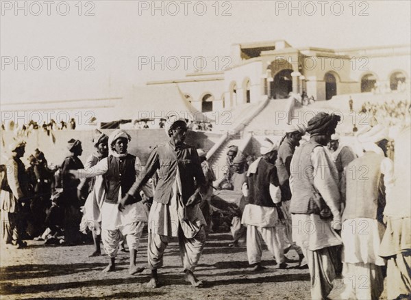 Male dancers in Sukkur. Male dancers perform to seated spectators at an outdoor event in Sukkur. Sukkur, Sind, India (Sindh, Pakistan), circa 1908. Sukkur, Sindh, Pakistan, Southern Asia, Asia.