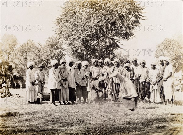 A wrestling match in Sukkur . Men in traditional Indian dress stand around a straw-covered patch of ground, watching as two men wrestle each other in a match. Sukkur, Sind, India (Sindh, Pakistan), circa 1908. Sukkur, Sindh, Pakistan, Southern Asia, Asia.