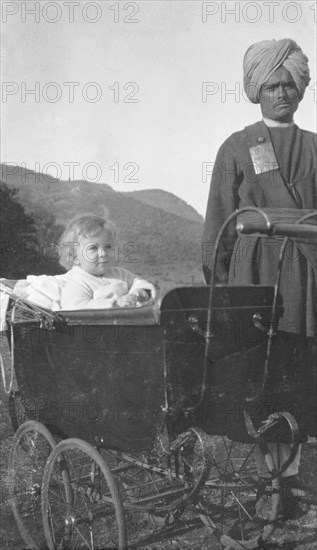 Indian servant with a British toddler. A turbaned Indian servant watches over a British toddler who sits upright in her pram outdoors. India, circa 1920. India, Southern Asia, Asia.