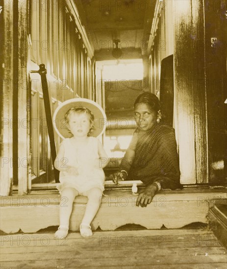Henry Lawrence with his ayah . Henry Lawrence (b.1902), the youngest son of Phyllis and Sir Henry Staveley Lawrence (a British civil servant), sits beside his Indian ayah (nursemaid) at the top of a flight of stairs wearing a large solatopi hat. India, circa 1905. India, Southern Asia, Asia.