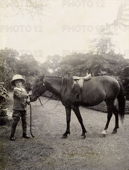 Owen with a pony, India. A European boy, Owen, wears a solatopi hat as he poses for the camera whilst holding the reigns of a saddled pony. Poona, Bombay Presidency (Pune, Maharashtra), India, circa 1920. Pune, Maharashtra, India, Southern Asia, Asia.